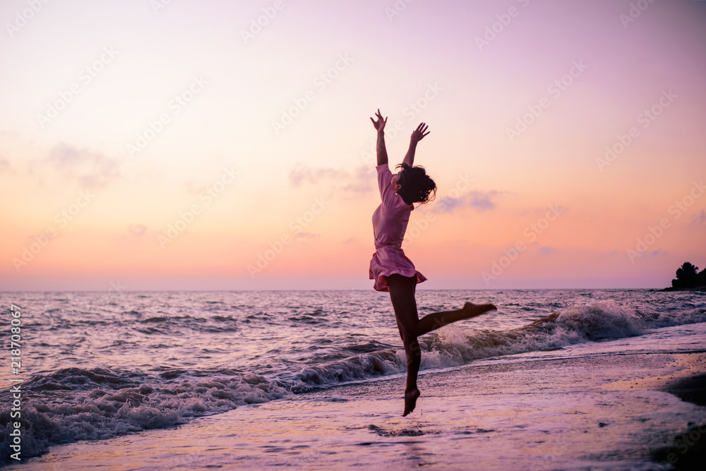 Silhouette of a stunning girl on the beach, on the background of a pink dawn, in the pose of a jumping ballerina on stage, with arms spread in the form of wings.