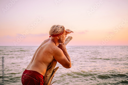 A tanned man stands on the coast leaning on an old tree, looking for a ship on the horizon that can pick him up from the island, after a shipwreck. photo