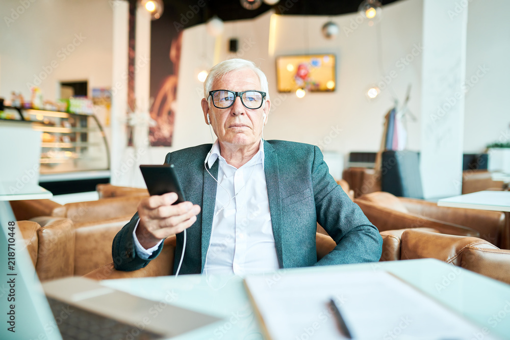 Portrait o modern senior businessman relaxing at table in cafe and listening to music from smartphone, copy space Portrait o modern senior businessman relaxing at table in cafe and listening to music from smartphone, copy space