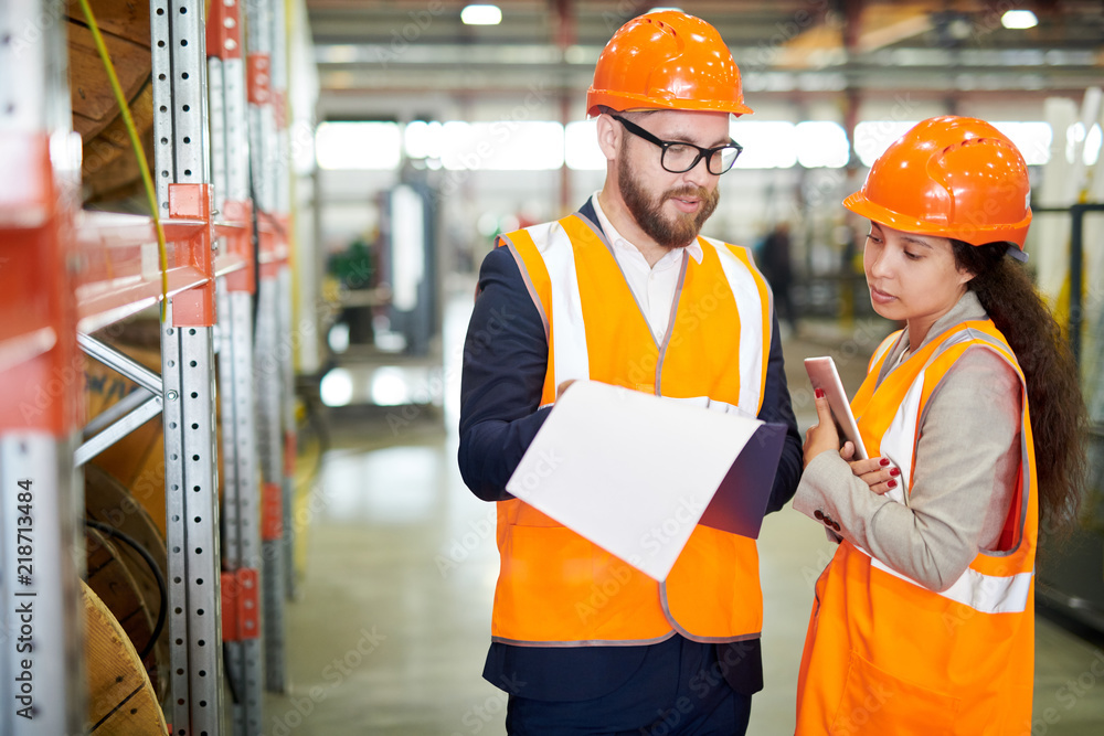 Portrait of mature businessman wearing hardhat talking to female factory worker standing by shelves in warehouse, copy space Portrait of mature businessman wearing hardhat talking to female factory worker standing by shelves in warehouse, copy space