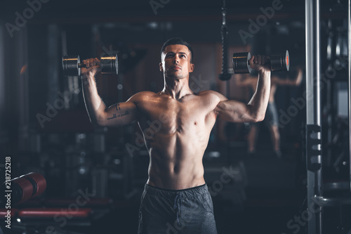 Shapely topless guy is standing and lifting dumbbells in gym. He is pressing equipment up with effort. Strong male training concept
