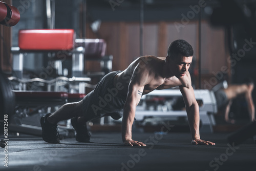 Concentrated ripped guy is exercising in sport center. He is staying in static position while doing plank. Shirtless male is balancing on feet and hands