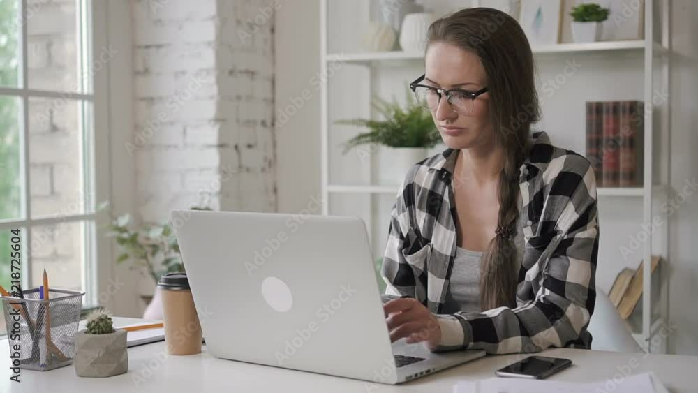 Female university saleswoman writing her course paper at home. Young woman with long hair sitting at the table with computer on, typing text, taking coffee cup from the desk and drinking. Working Female university saleswoman writing her course paper at home. Young woman with long hair sitting at the table with computer on, typing text, taking coffee cup from the desk and drinking. Working