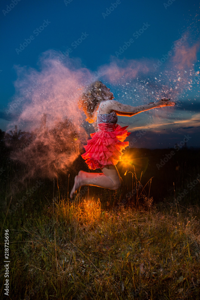 Obraz premium A girl in a black top and red skirt jumping in a field during photoshoot with flour and sunset behing her