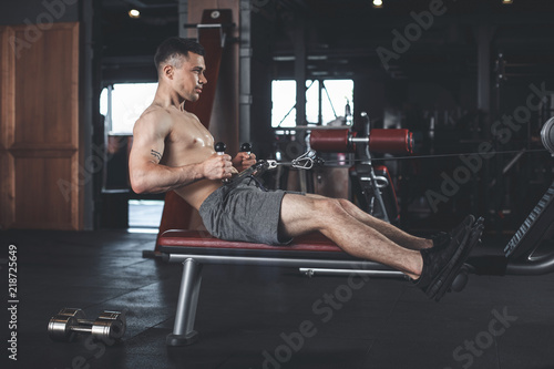 Man is training with row equipment by pulling handles while sitting. He is exercising back muscles with weight. Guy is using different outfit during active time in gym