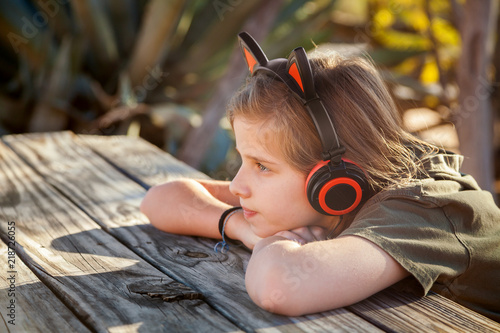 Young Blond Girl Wearing Earphone With Cat Ears on Top Sitting Calmly at Picnic Table Outside in the Desert While the Sun Sets photo