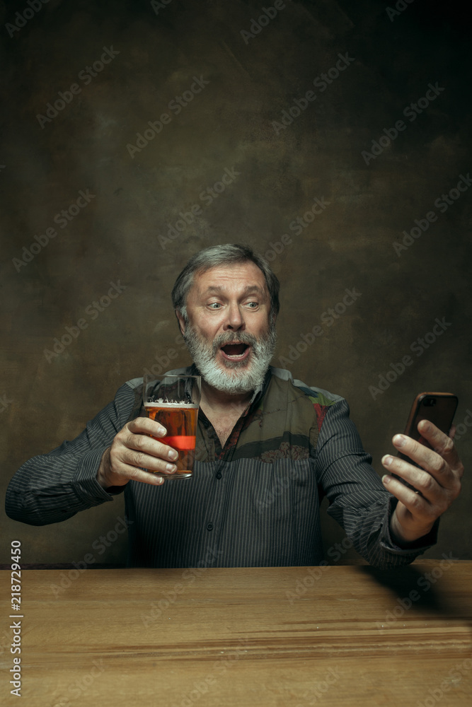Smiling bearded man drinking alcohol in pub. Enjoying my favorite beer. The front view of handsome smiling senior man with glass of beer sitting at the wooden table. Studio shot with caucasian model Smiling bearded man drinking alcohol in pub. Enjoying my favorite beer. The front view of handsome smiling senior man with glass of beer sitting at the wooden table. Studio shot with caucasian model