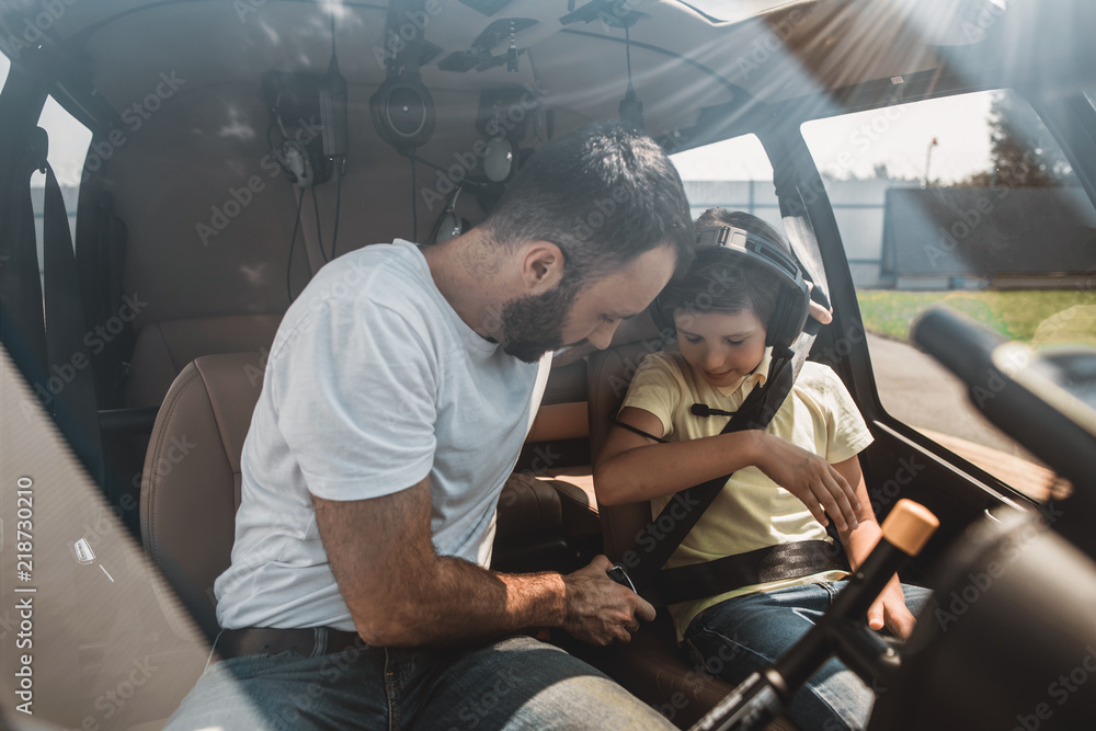 Orderly bearded male fastening seat belt on positive kid while telling with him inside of rotorcraft. Outgoing child looking at it and gesticulating hand. He putting headset on Orderly bearded male fastening seat belt on positive kid while telling with him inside of rotorcraft. Outgoing child looking at it and gesticulating hand. He putting headset on