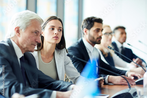 Young female delegate in elegant suit listening to speaker very attentively among her foreign colleagues photo