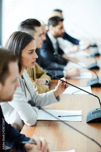 Young brunette female in formalwear speaking in microphone at conference or summit among her colleagues photo