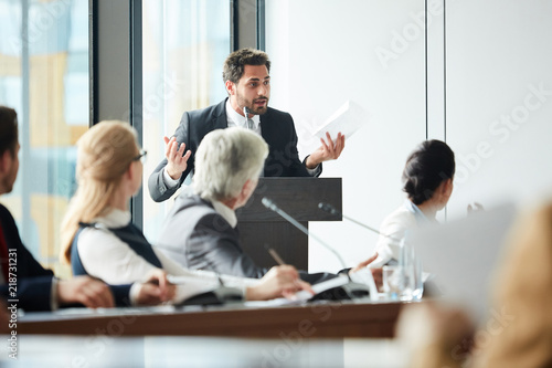 Young delegate with paper speaking in microphone by tribune while debating with opponent