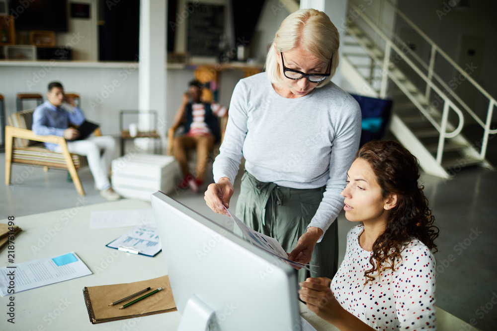 Mature female leader wearing eyeglasses discussing documents with businesswoman while she is sitting at the desk Mature female leader wearing eyeglasses discussing documents with businesswoman while she is sitting at the desk