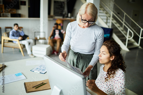 Mature female leader wearing eyeglasses discussing documents with businesswoman while she is sitting at the desk