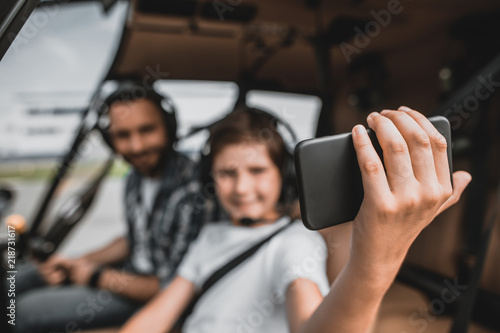 Portrait of glad kid keeping contemporary mobile in hand while doing felfie with positive dad. They having fun in rotor plane