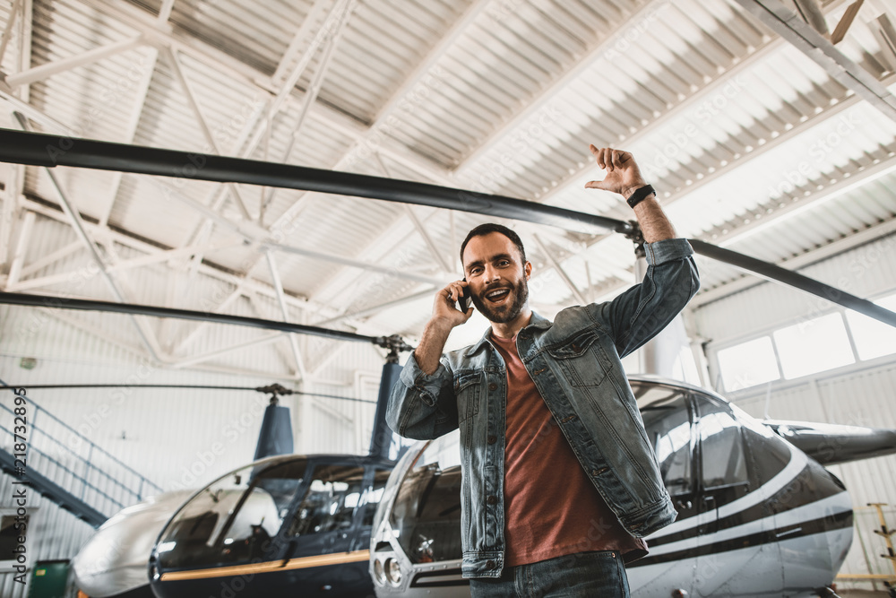 Low angle portrait of bearded cheerful male worker talking on cellphone while gesticulating hands. He locating indoor near rotor plane Low angle portrait of bearded cheerful male worker talking on cellphone while gesticulating hands. He locating indoor near rotor plane