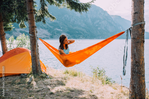Woman traveker relaxing in a hammock overlooking the water under trees pine enjoying the view at the lake in summer norwegian cloudy morning. Rest on camping concept.