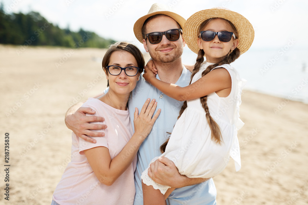 Affectionate family of father, mother and little daughter embracing while enjoying summer weekend on the beach Affectionate family of father, mother and little daughter embracing while enjoying summer weekend on the beach