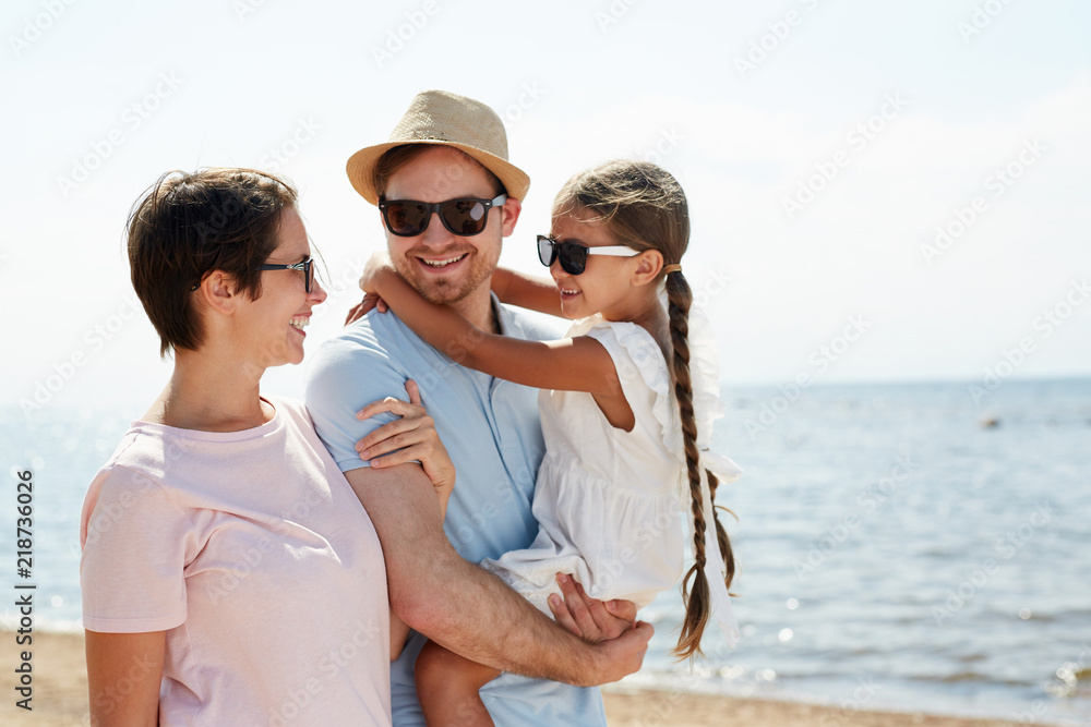 Happy young family consisting of father, mother and cute little daughter laughing and enjoying chill by seaside Happy young family consisting of father, mother and cute little daughter laughing and enjoying chill by seaside