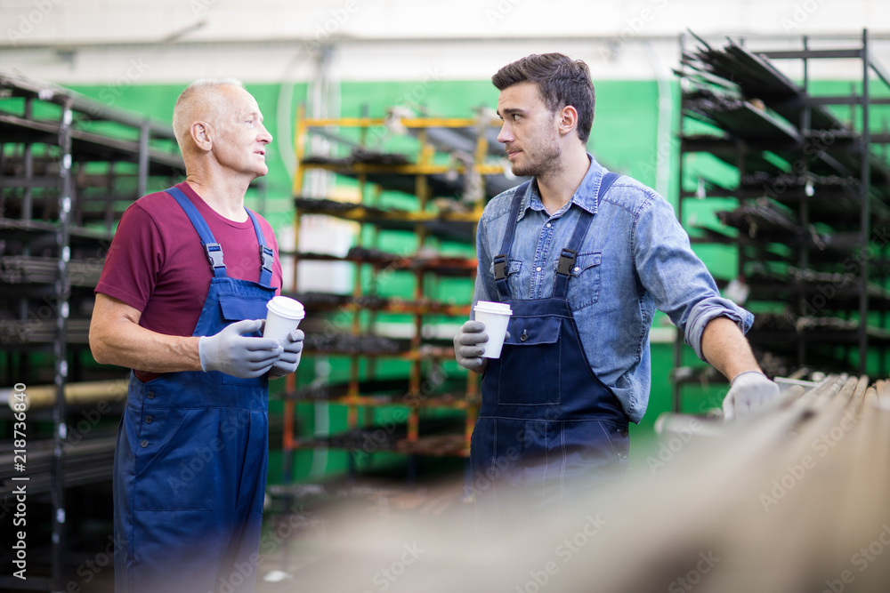 Two colleagues standing in overalls taking coffee break and communicating in workshop Two colleagues standing in overalls taking coffee break and communicating in workshop