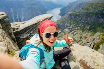 Sportive smiling woman hiker in eyeglasses making selfie portrait above Trolltunga and Norwegian fjord, Norway. Adventure, travel and hiking concept.