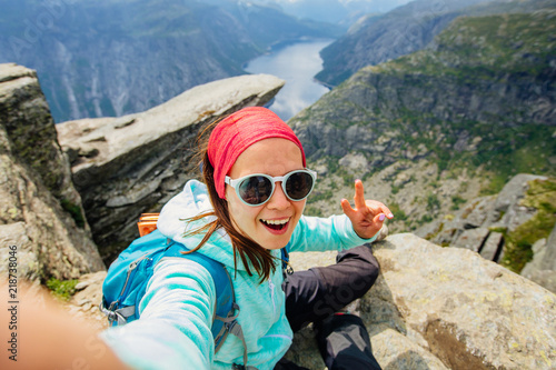 Sportive smiling woman hiker in eyeglasses making selfie portrait above Trolltunga and Norwegian fjord, Norway. Adventure, travel and hiking concept.