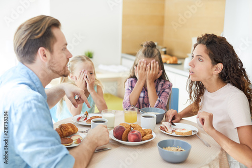 Young annoyed couple having argument by breakfast while their daughters sitting near by and covering faces by hands