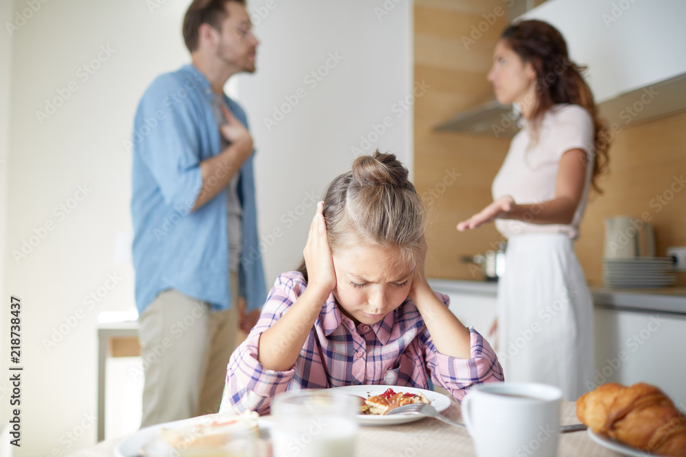Stressed girl covering her ears while leaning over served table at breakfast on background of her parents quarreling Stressed girl covering her ears while leaning over served table at breakfast on background of her parents quarreling