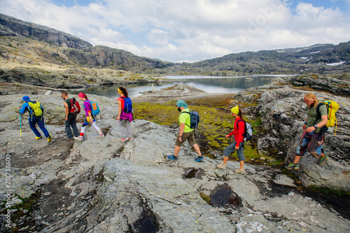 Group of seven hikers men and woman walking with backpacks and trekking poles along water lake in mountain valley during Norway travel. photo