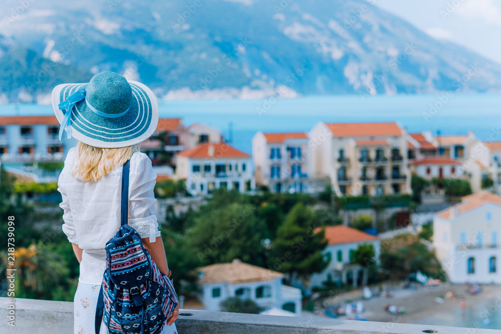 Young stylish traveler woman enjoying view of colorful tranquil Assos village. Female model wearing blue sunhat, white clothes and travel backpack enjoying her summer vacation in Kefalonia, Greece