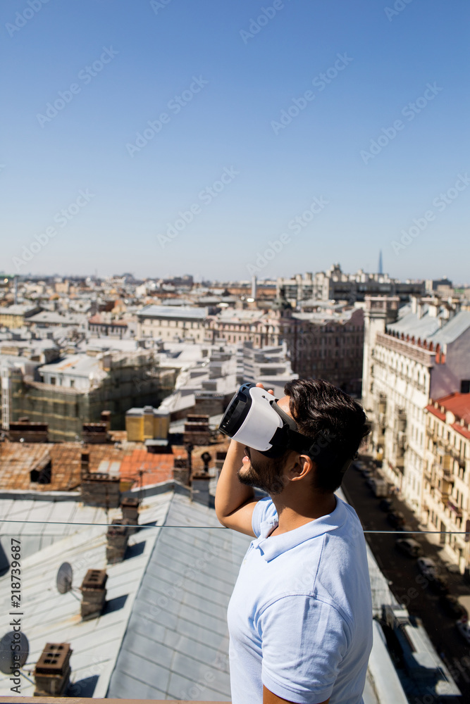 Young modern man in vr headset looking at sun on clear blue sky with cityscape on background