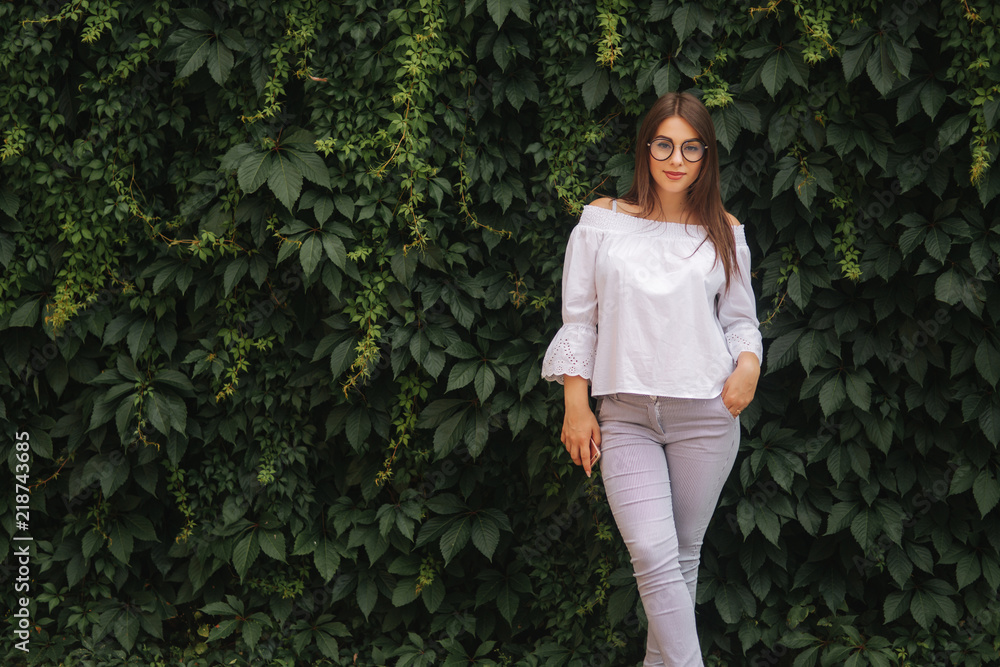 Girl girl smile to camera. Female is dressed in a white shirt is standing near a wall overgrown with ivy Girl girl smile to camera. Female is dressed in a white shirt is standing near a wall overgrown with ivy