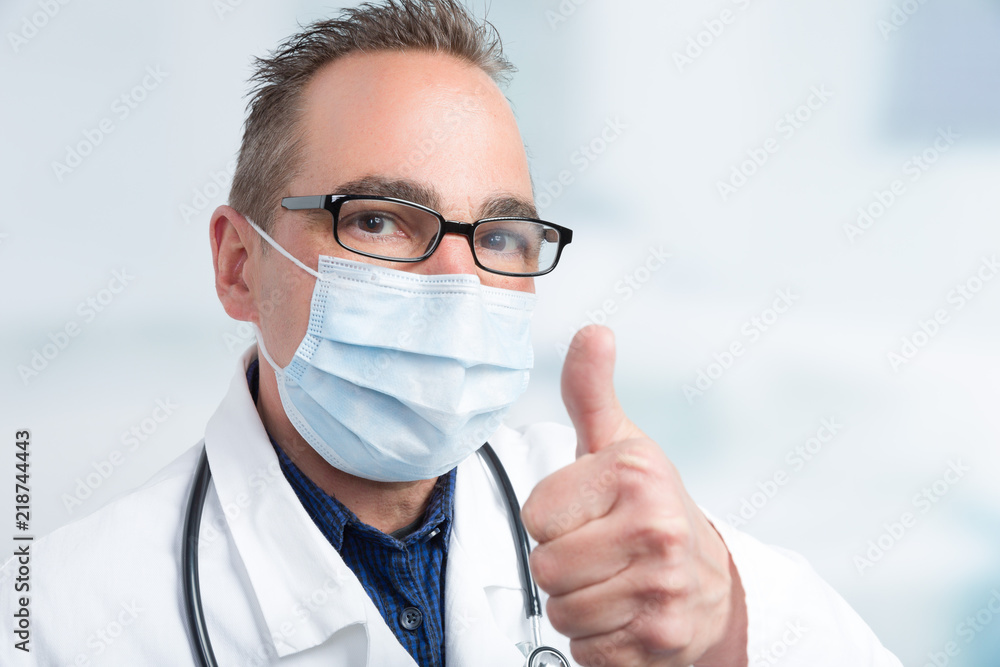 Ein Arzt mit Mundschutz mit Klinikraum im iIntergrund zeigt Daumen hoch Ein Arzt mit Mundschutz mit Klinikraum im iIntergrund zeigt Daumen hoch