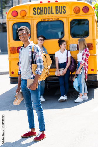 handsome smiling teen schoolboy looking at camera while his classmates standing behind near school bus