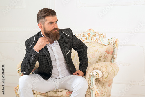 Imposing well dressed man sitting on couch. Men's beauty, fashion. Cool man in formal wear, tux pocket square, sit in chair over white background, looking away, shares, stock, money, rich, brutal.