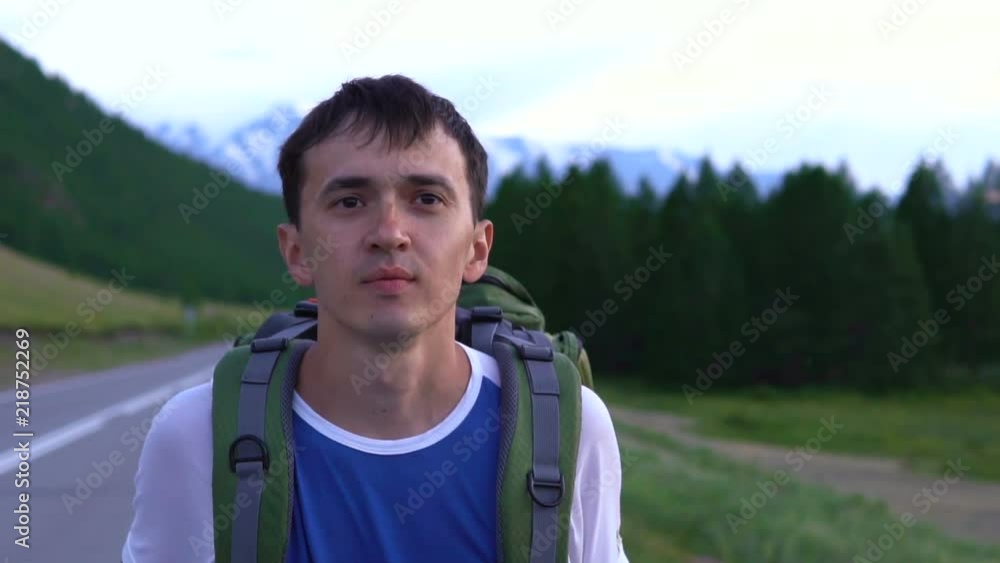 A tourist hitchhiker walks along the asphalt road with a backpack on his shoulders. Ahead is a snowy peak of the mountain. A tourist hitchhiker walks along the asphalt road with a backpack on his shoulders. Ahead is a snowy peak of the mountain.