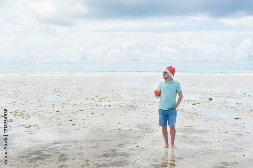 A man with a beard in a Christmas hat drinks a cocktail on the beach of Zanzibar island. New year's eve in an exotic country