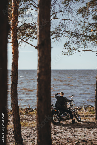 Fototapeta Naklejka Na Ścianę i Meble -  biker in black leather jacket sitting on classical chopper motorcycle near river in forest