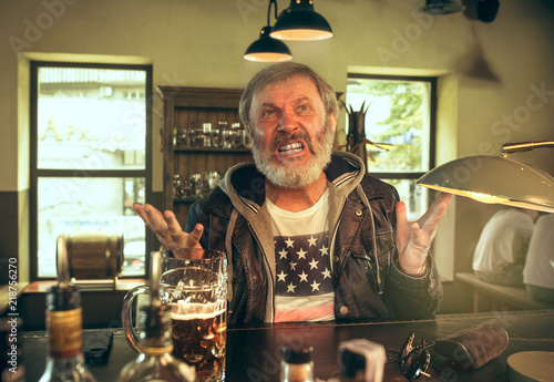 Angry bearded man drinking alcohol in pub and watching a sport program on TV. Enjoying my favorite teem and beer. Man with mug of beer sitting at table. Football or sport fan. Human emotions concept