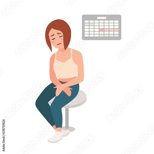 Sad girl sitting on stool with her hands on belly, suffering from menstrual pain and cramps, crying against calendar hanging on wall on background