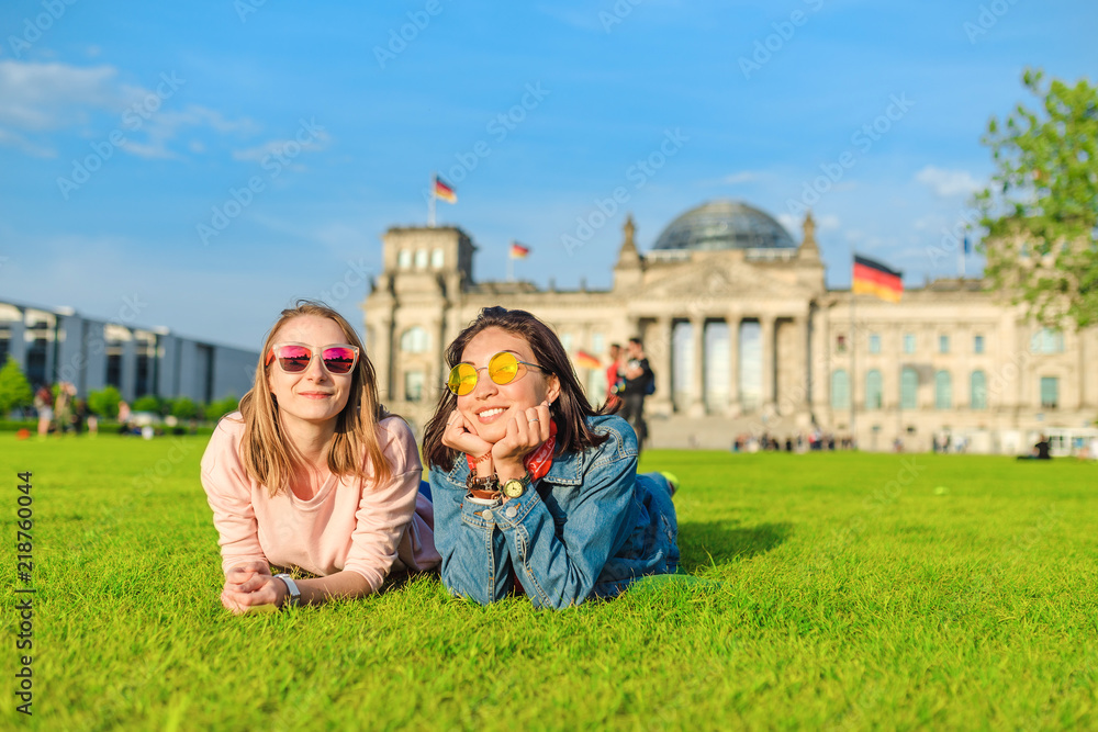 Obraz premium Dwie młode szczęśliwe dziewczyny noszące okulary przeciwsłoneczne leżące na trawie i bawią się przed budynkiem Bundestagu w Berlinie. Studiowanie za granicą i podróżowanie po Niemczech