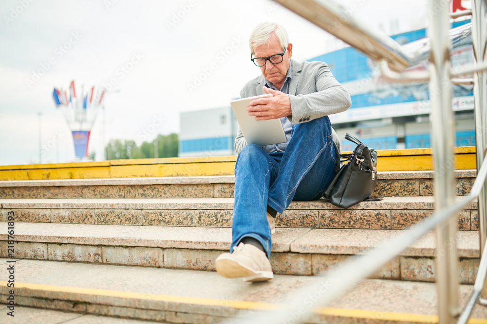 Senior man in glasses sitting on street stairs near bag and browsing modern tablet Senior man in glasses sitting on street stairs near bag and browsing modern tablet