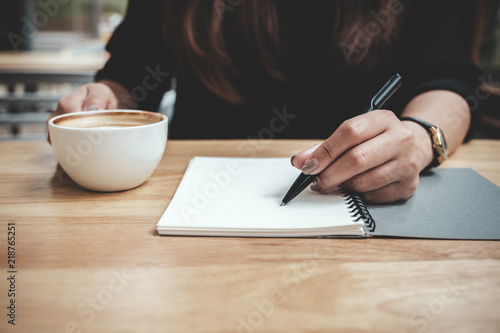 Closeup image of a woman's hand writing down on a white blank notebook while drinking coffee on wooden table