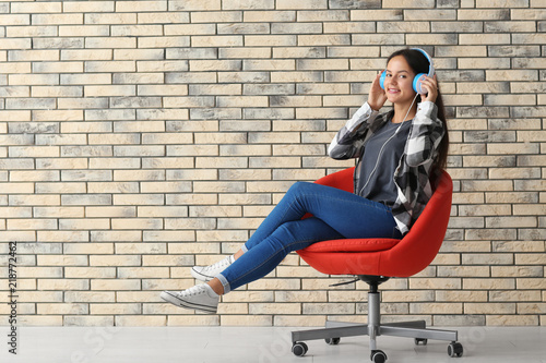 Beautiful young woman with headphones listening to music while sitting on armchair near brick wall © Pixel-Shot