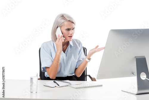 portrait of businesswoman talking on smartphone at workplace with glass of water, notebook and computer screen isolated on white