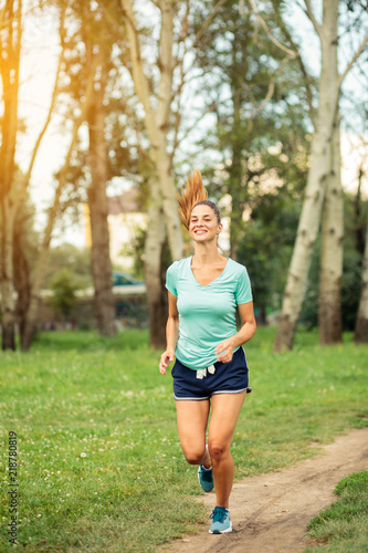 Happy young woman running along the footpath in a city park. Jogging and exercising in urban environment. Sport and fitness concept