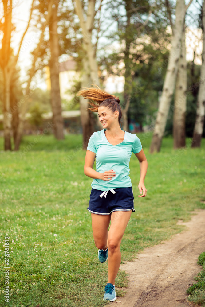 Joyful young woman jogging along the footpath in a city park. Running and exercising in urban environment. Sport and healthy lifestyle concept Joyful young woman jogging along the footpath in a city park. Running and exercising in urban environment. Sport and healthy lifestyle concept