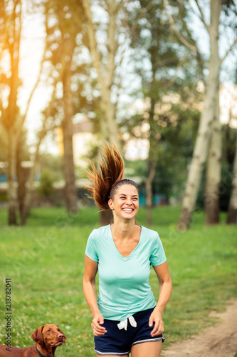 Smiling happy young woman running with her pet dog through a city park. Jogging and exercising in urban environment. Sport and healthy lifestyle concept