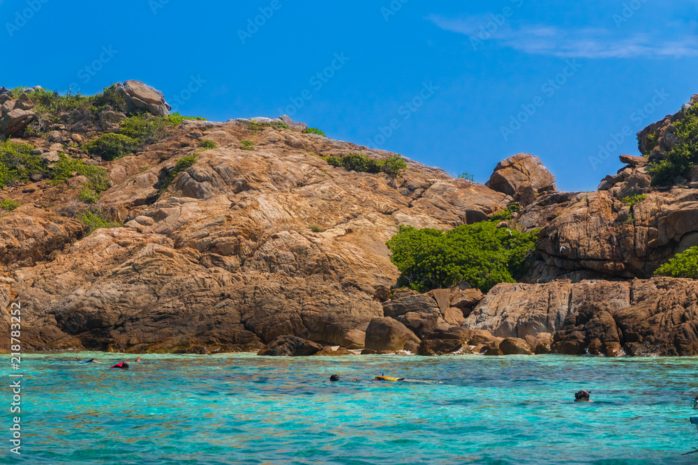 Fototapeta premium Lovely scenery with a blue sky, turquoise water & tourists snorkeling in front of the towering boulder rocks of Tokong Burung (Bird Island), a popular snorkeling spot near Perhentian Kecil, Malaysia.