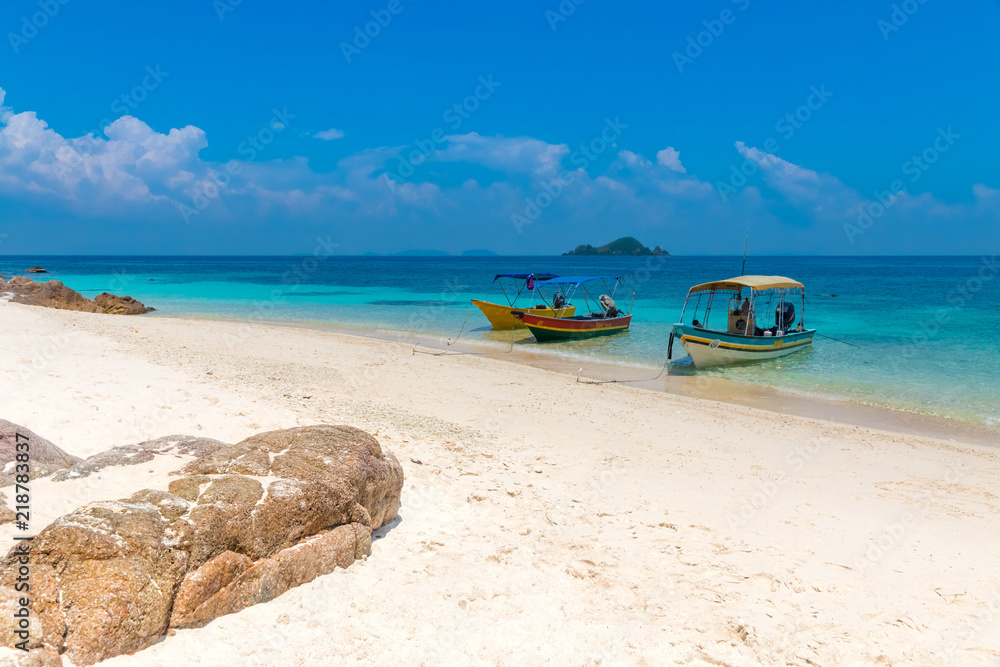 Three tourist boats anchoring on a white powdery sandy beach, a blue sky and a small island on the horizon makes it a gorgeous scene of the uninhabited Rawa Island near Perhentian Kecil in Malaysia.