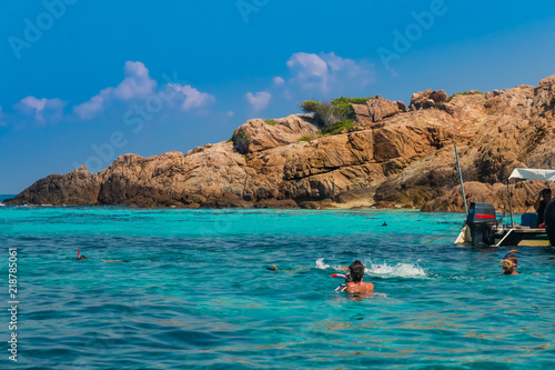 Next to a boat, tourists are snorkeling in crystal clear turquoise blue water in front of the magnificent rocks of Tokong Burung (Bird Island), an uninhabited island near Perhentian Kecil, Malaysia. 